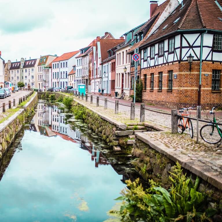 Home 3 Ein schmaler, von Steinmauern gesäumter Kanal verläuft entlang einer kopfsteingepflasterten Straße mit bunten, historischen europäischen Häusern, die an den Charme der schönsten Städte Deutschlands erinnern. Zwei Fahrräder stehen am Wasser, eingerahmt von üppigen Pflanzen und einem wolkenverhangenen Himmel, der sich darunter spiegelt.