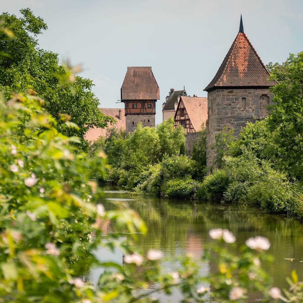 Home 7 Eine malerische Ansicht eines mittelalterlichen Steinturms mit Spitzdach in Dinkelsbühl, umgeben von üppigen grünen Bäumen und Sträuchern. Im Vordergrund fließt ein ruhiger Fluss, in dem sich das Grün und die historischen Gebäude mit steilen Dächern im Hintergrund spiegeln.