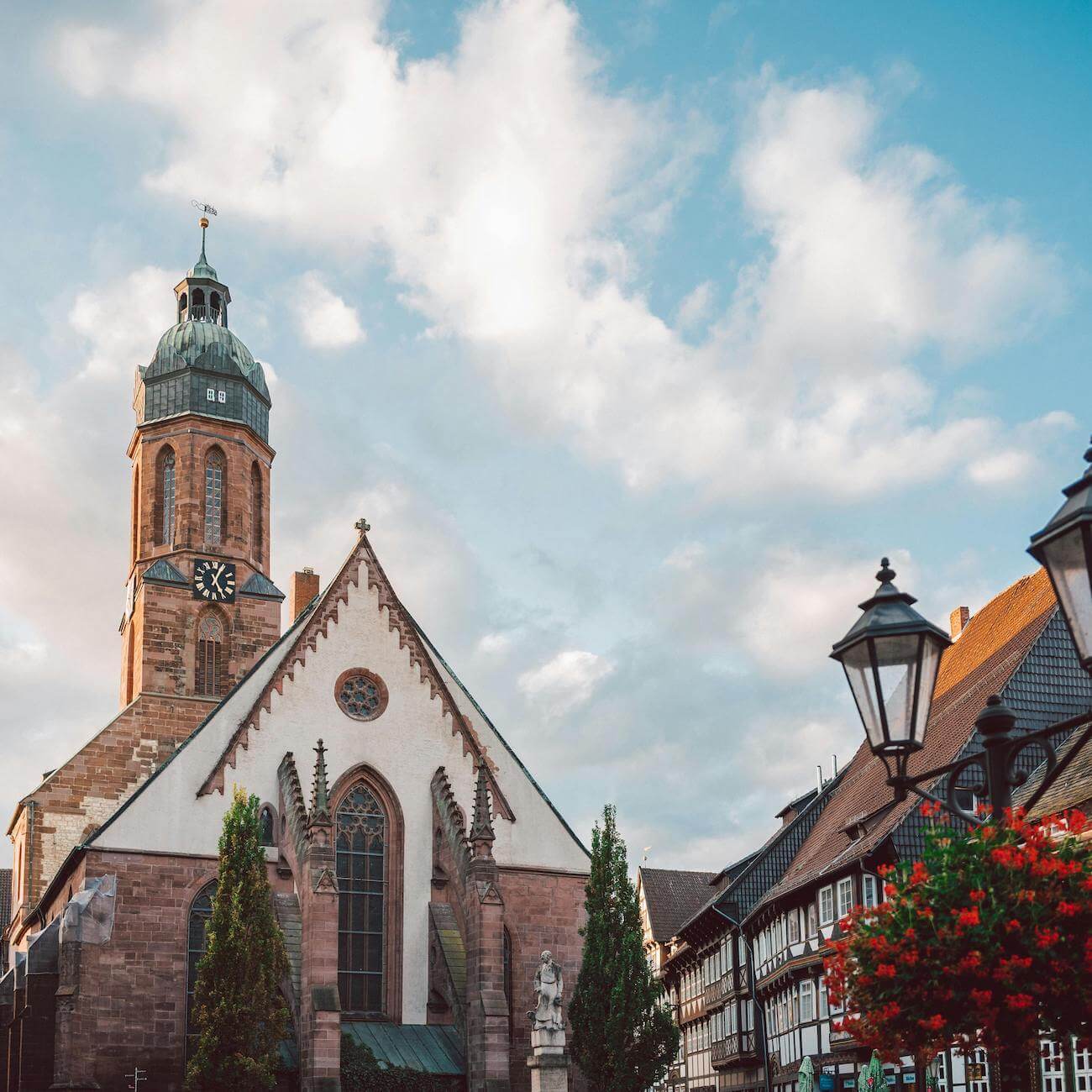 Home 2 Eine historische Kirche mit einem hohen Glockenturm und gewölbten Fenstern steht unter einem teilweise bewölkten Himmel. Traditionelle Fachwerkgebäude und altmodische Straßenlaternen umgeben die Kirche. An einem Blumenkübel im Vordergrund hängen leuchtend rote Blumen.