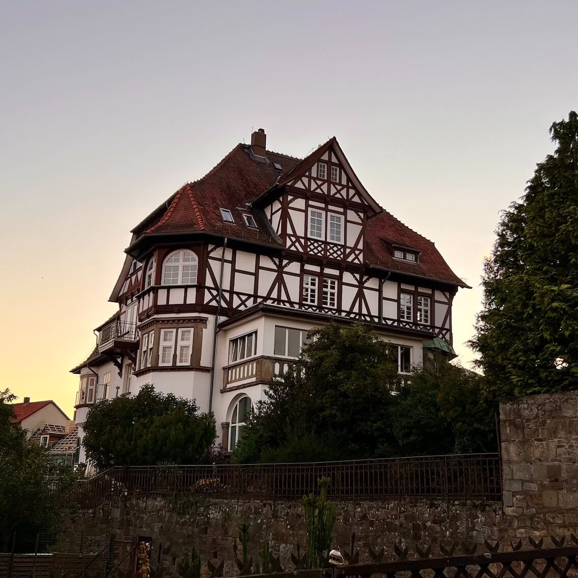 Home 5 Ein großes, traditionelles deutsches Fachwerkhaus mit roten Ziegeldächern, weißen Wänden und dunklen Holzbalken steht hinter einem Steinzaun, umgeben von Grün, unter einem klaren Abendhimmel mit weichem, warmem Licht.