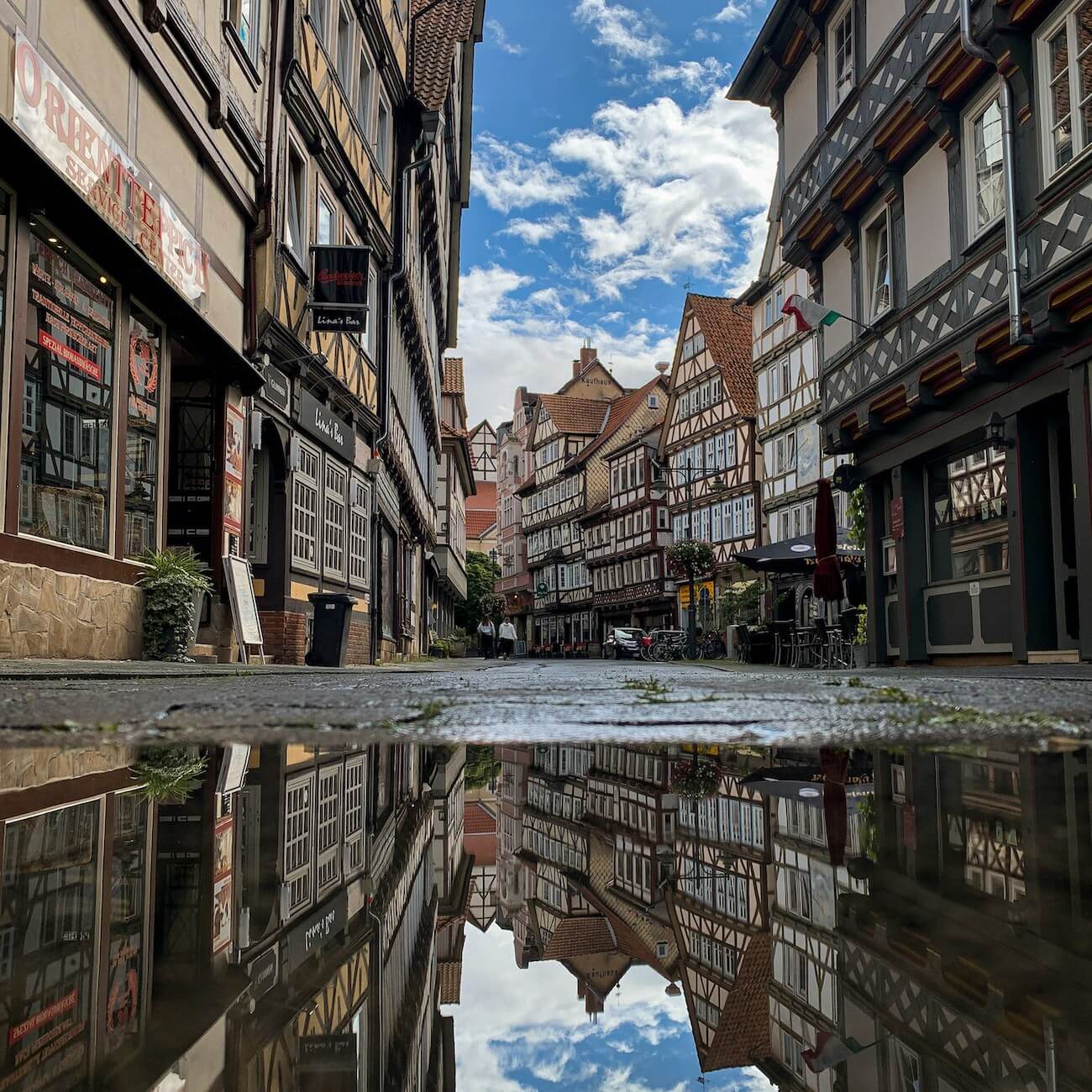 Home 4 Eine Kopfsteinpflasterstraße in Hannoversch Münden, gesäumt von alten Fachwerkhäusern, spiegelt sich in einer großen Pfütze. Der blaue, teilweise wolkenverhangene Himmel und die historischen Dächer spiegeln sich im Wasser und erzeugen einen auffallend symmetrischen Effekt.
