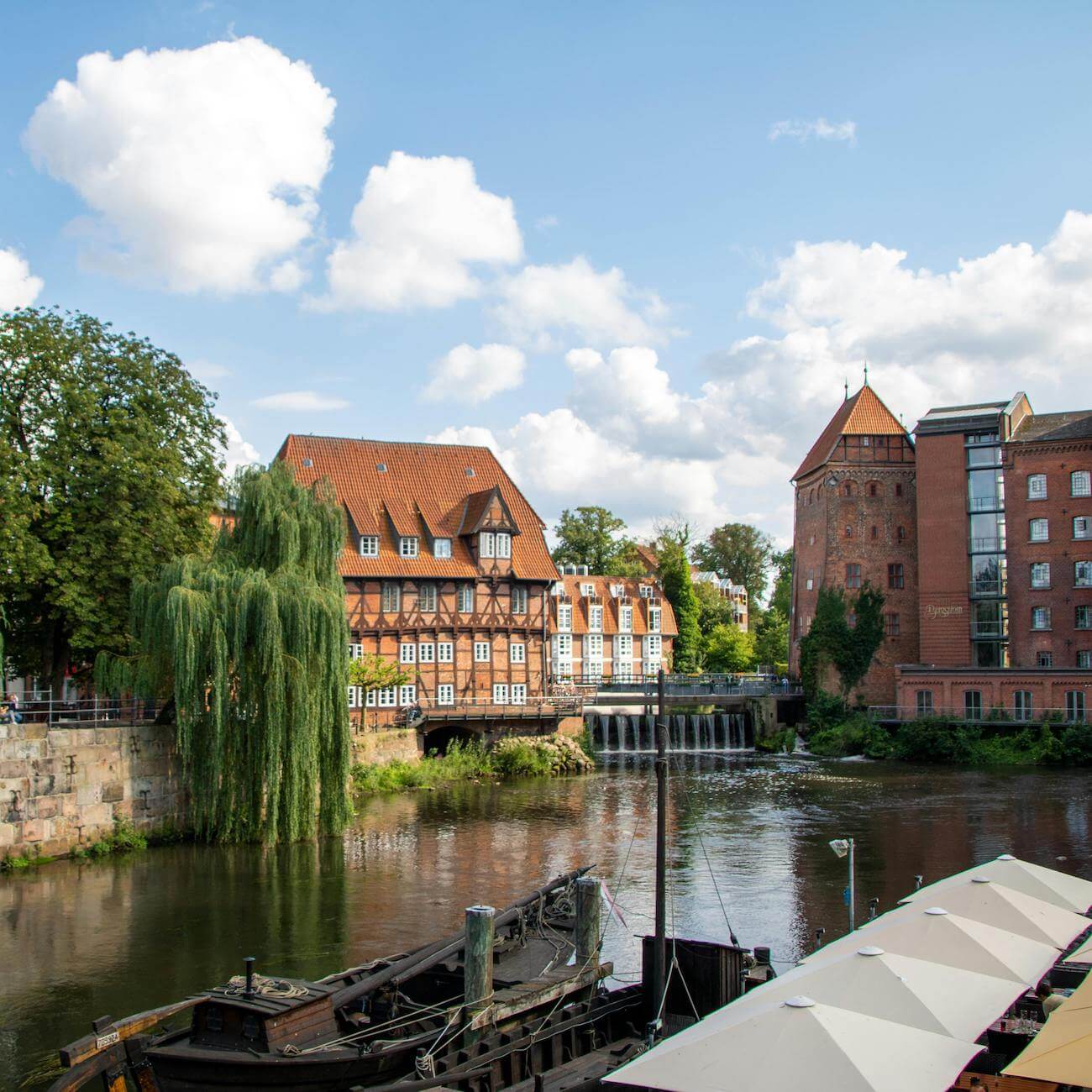 Home 1 Eine malerische Flusslandschaft in Lüneburg zeigt historische Backsteingebäude, darunter ein Fachwerkhaus und einen Turm, entlang eines ruhigen Flusses. Ein altes Holzboot liegt in der Nähe des Ufers, während Bäume und Wolken die friedliche, sonnige Szene vervollständigen.