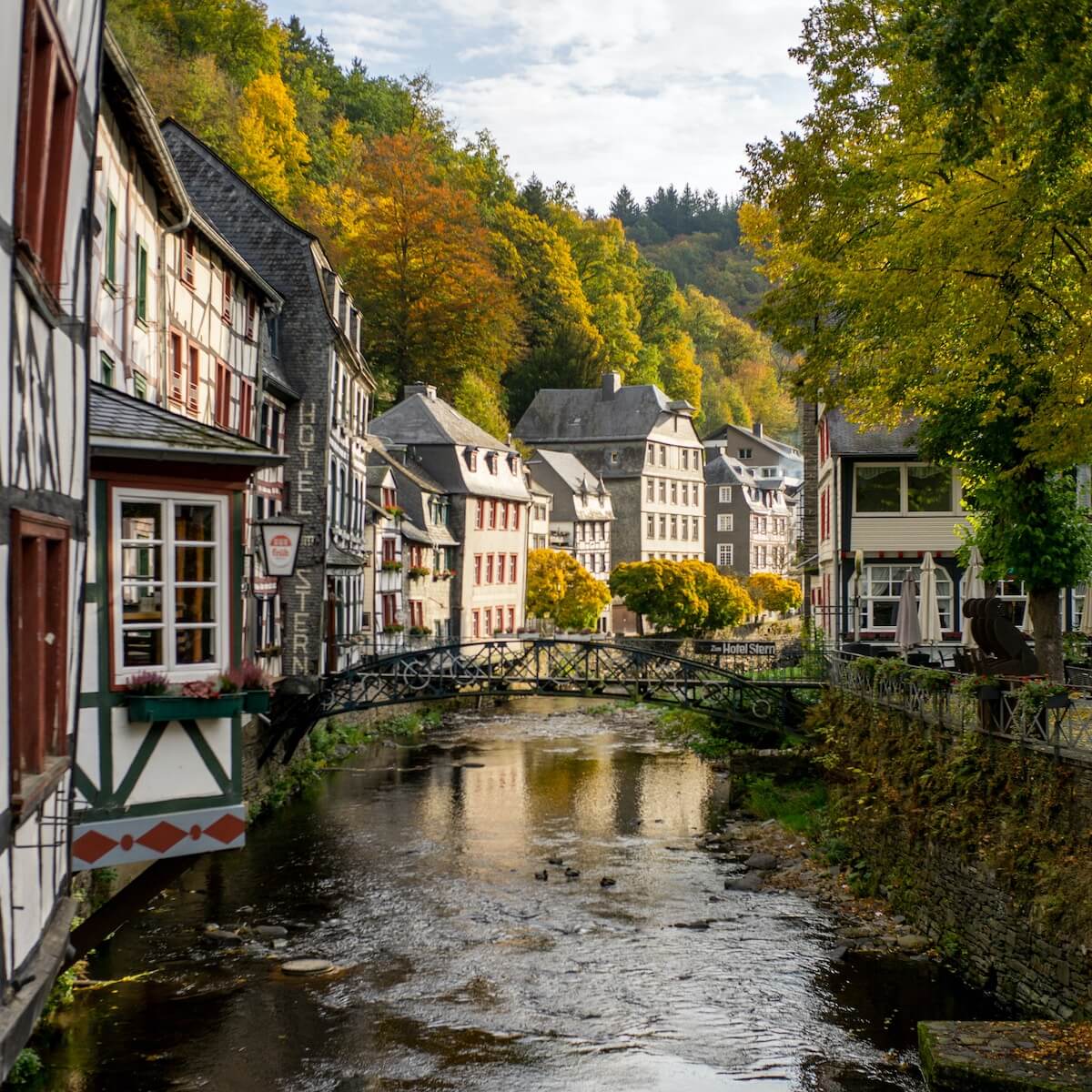 Home 2 Ein schmaler Fluss fließt durch das malerische europäische Dorf Monschau, an dessen beiden Ufern Fachwerkhäuser stehen. Eine kleine Eisenbrücke überquert das Wasser, umgeben von Bäumen mit grünen und goldenen Herbstblättern unter einem teilweise bewölkten Himmel.