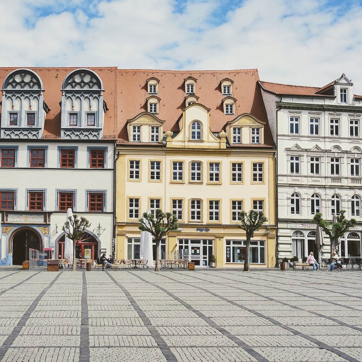 Home 4 Ein breiter kopfsteingepflasterter Platz in Naumburg führt zu farbenfrohen, historischen europäischen Gebäuden mit verzierten Fassaden und roten Ziegeldächern. Laublose, beschnittene Bäume säumen den Platz, während sich die Menschen an Tischen im Freien in der Nähe des blassgelben Hauptgebäudes unter einem teilweise bewölkten Himmel entspannen.