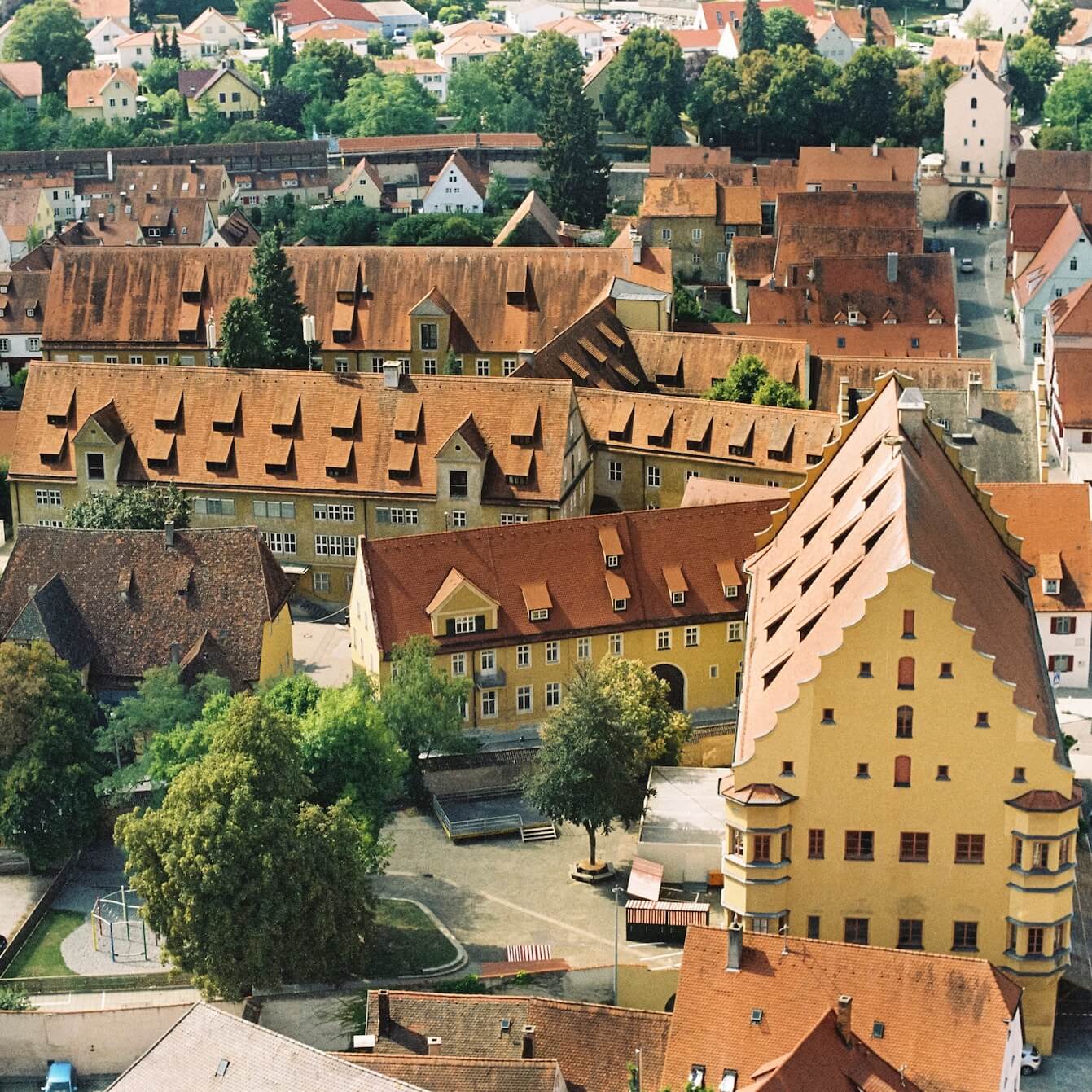 Home 5 Luftaufnahme von Nördlingen, einer europäischen Stadt mit roten Ziegeldächern, gelben und beigen Gebäuden und Giebelfassaden. Zu sehen sind Bäume und ein kleiner Innenhof, ein Spielplatz und schmale Straßen, die sich durch dicht gedrängte Häuser schlängeln.