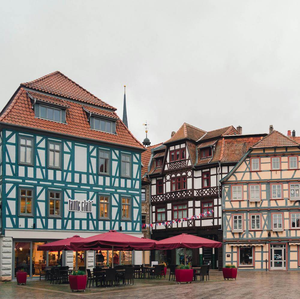 Home 5 Ein Stadtplatz mit bunten Fachwerkhäusern, Sitzgelegenheiten im Freien unter kastanienbraunen Sonnenschirmen, Topfpflanzen und einem bewölkten Himmel. Auf dem linken Gebäude ist das "Café Chilli" zu sehen; die Architektur zeichnet sich durch verschnörkelte Holzarbeiten und steile Dächer aus.