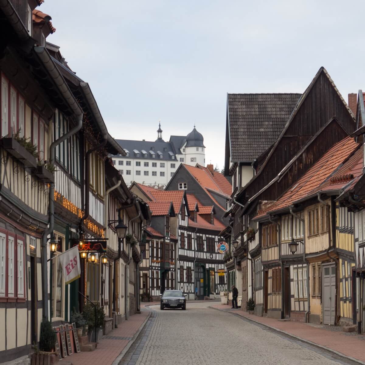 Home 3 Eine schmale Straße mit Kopfsteinpflaster, gesäumt von alten Fachwerkhäusern, von denen einige mit warmen Lichtern beleuchtet sind. Am Ende der Straße erhebt sich ein großes weißes, schlossähnliches Gebäude über den Dächern unter einem bewölkten Himmel. Ein einzelnes geparktes Auto und eine Person sind zu sehen.