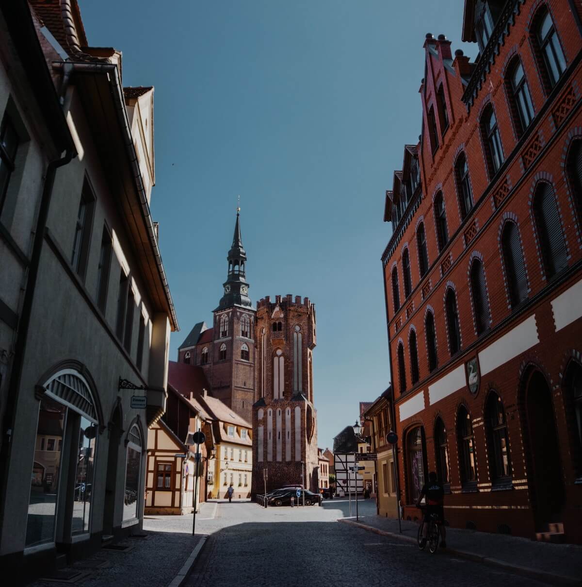 Home 7 Eine Kopfsteinpflasterstraße in Tangermünde, die von alten, verzierten Gebäuden gesäumt ist, führt zu einer großen, historischen Kirche mit einer hohen Turmspitze und einem Glockenturm unter einem klaren blauen Himmel. Eine Person fährt mit dem Fahrrad in der Nähe des roten Backsteingebäudes, während sich die Schatten über die ruhige Straße erstrecken.