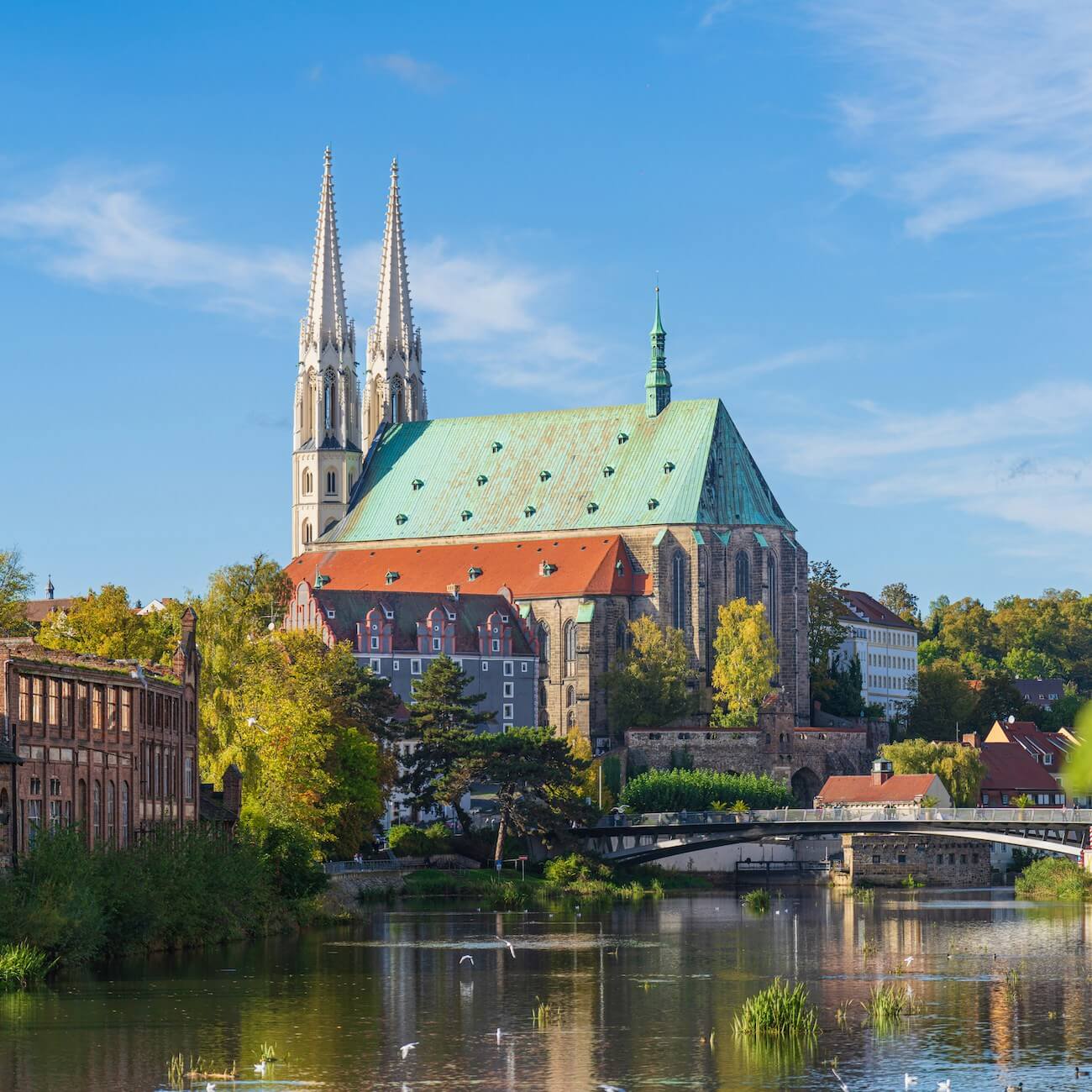 Home 6 Eine große gotische Kirche mit zwei hohen Türmen und einem grünen Kupferdach steht an einem Fluss in Görlitz. Sie ist von roten Dächern und Bäumen umgeben, und eine Brücke führt über das Wasser. Blauer Himmel und vereinzelte Wolken vervollständigen die ruhige, malerische Szene.