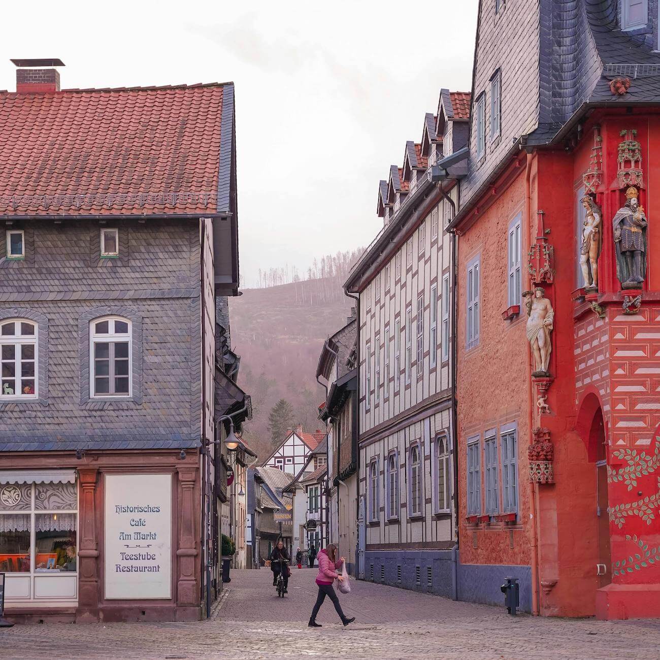 Home 9 Eine Kopfsteinpflasterstraße in Goslar schlängelt sich zwischen alten europäischen Gebäuden mit bunten Fassaden und kunstvollen Statuen. Eine Frau in einer rosafarbenen Jacke geht über die Straße, während sich zwei Radfahrer nähern. In der dunstigen Ferne erheben sich Hügel und kahle Bäume.