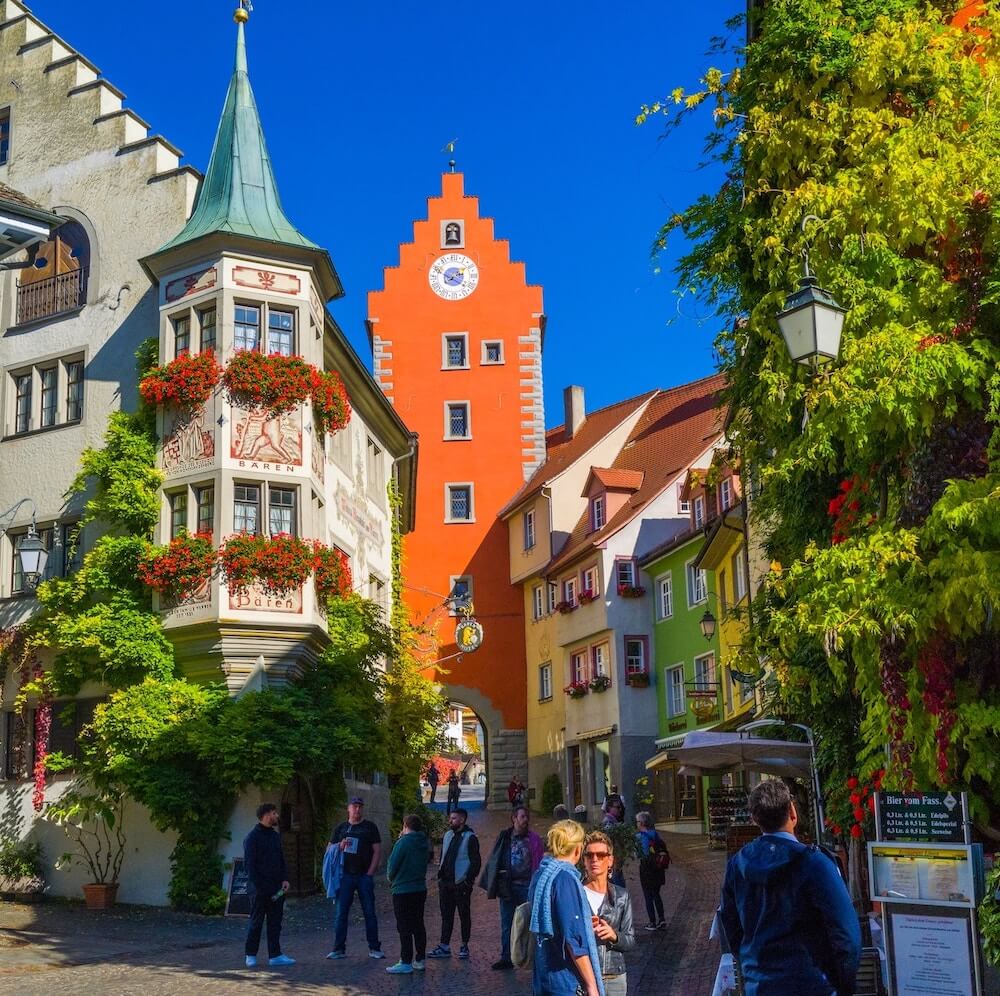Home 1 Eine lebhafte europäische Straßenszene in Meersburg mit farbenfrohen Gebäuden, darunter ein hoher orangefarbener Turm mit einer Uhr. Blumenkästen schmücken die Fenster, Grün klettert an den Fassaden empor, und die Menschen schlendern unter blauem Himmel die Kopfsteinpflasterstraße entlang.