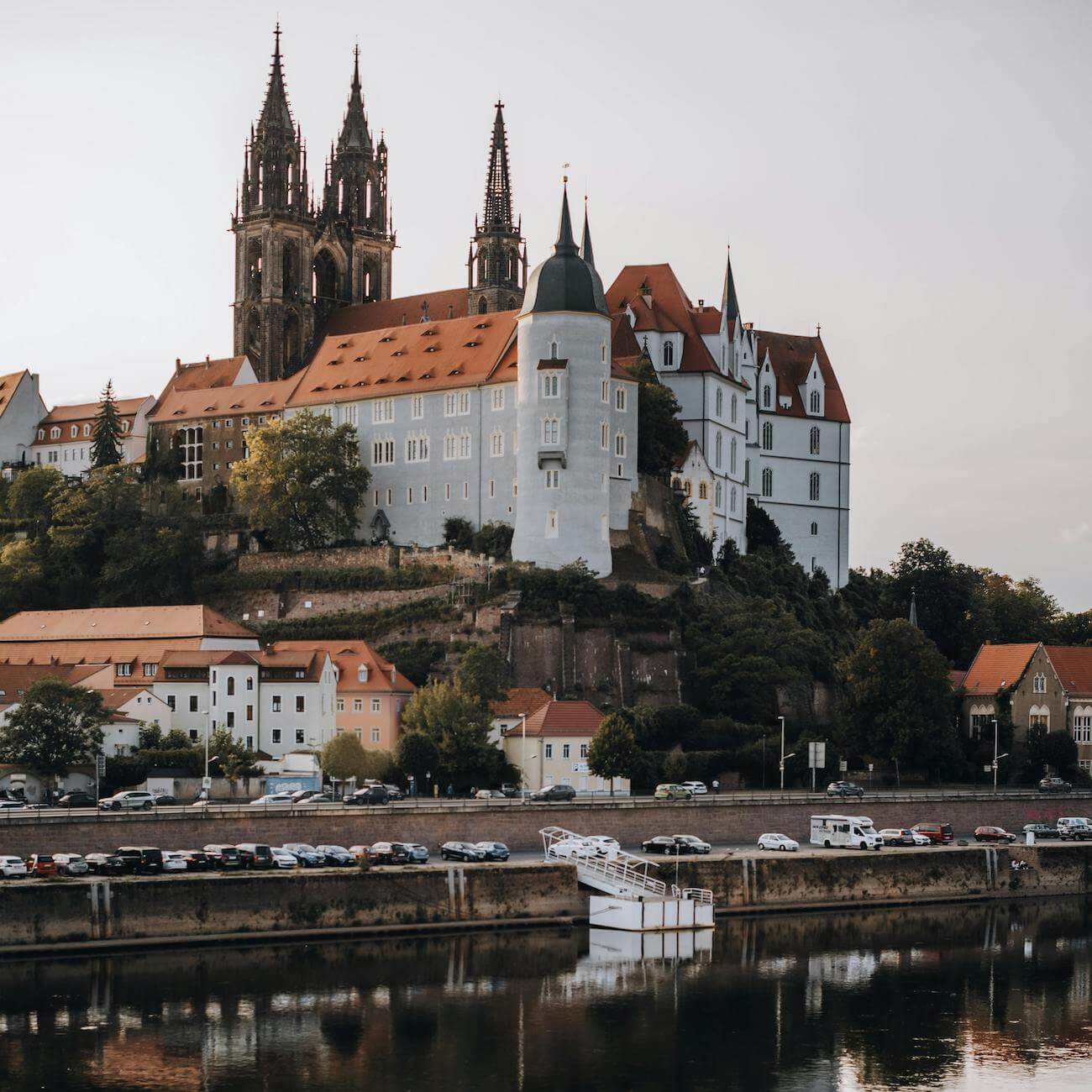 Home 1 Ein großes historisches Schloss mit roten Dächern und spitzen Türmen steht auf einem Hügel in Meißen, umgeben von Bäumen und kleineren Gebäuden. Im Vordergrund befindet sich ein Fluss mit geparkten Autos entlang des Ufers, in dessen ruhigem Wasser sich das Schloss und der Himmel spiegeln.