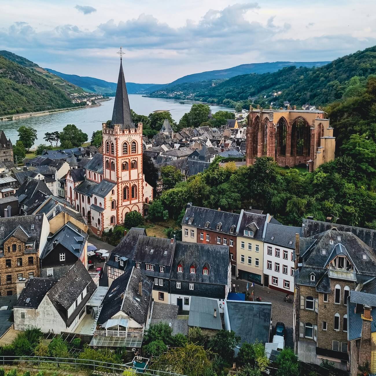 Home 8 Luftaufnahme des malerischen europäischen Dorfs Bacharach an einem Fluss, mit einer hohen Kirche mit spitzem Turm, historischen Stein- und Fachwerkhäusern, grünen Hügeln im Hintergrund und Ruinen eines alten Gebäudes in der Nähe der Kirche.