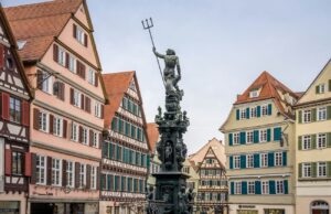 Auf einem kopfsteingepflasterten Platz in Tübingen, umgeben von bunten Fachwerkhäusern mit steilen roten Ziegeldächern und vielen Fenstern, steht eine Statue von Neptun mit einem Dreizack auf einem kunstvollen Brunnen und versprüht bei bedecktem Himmel europäischen mittelalterlichen Charme.
