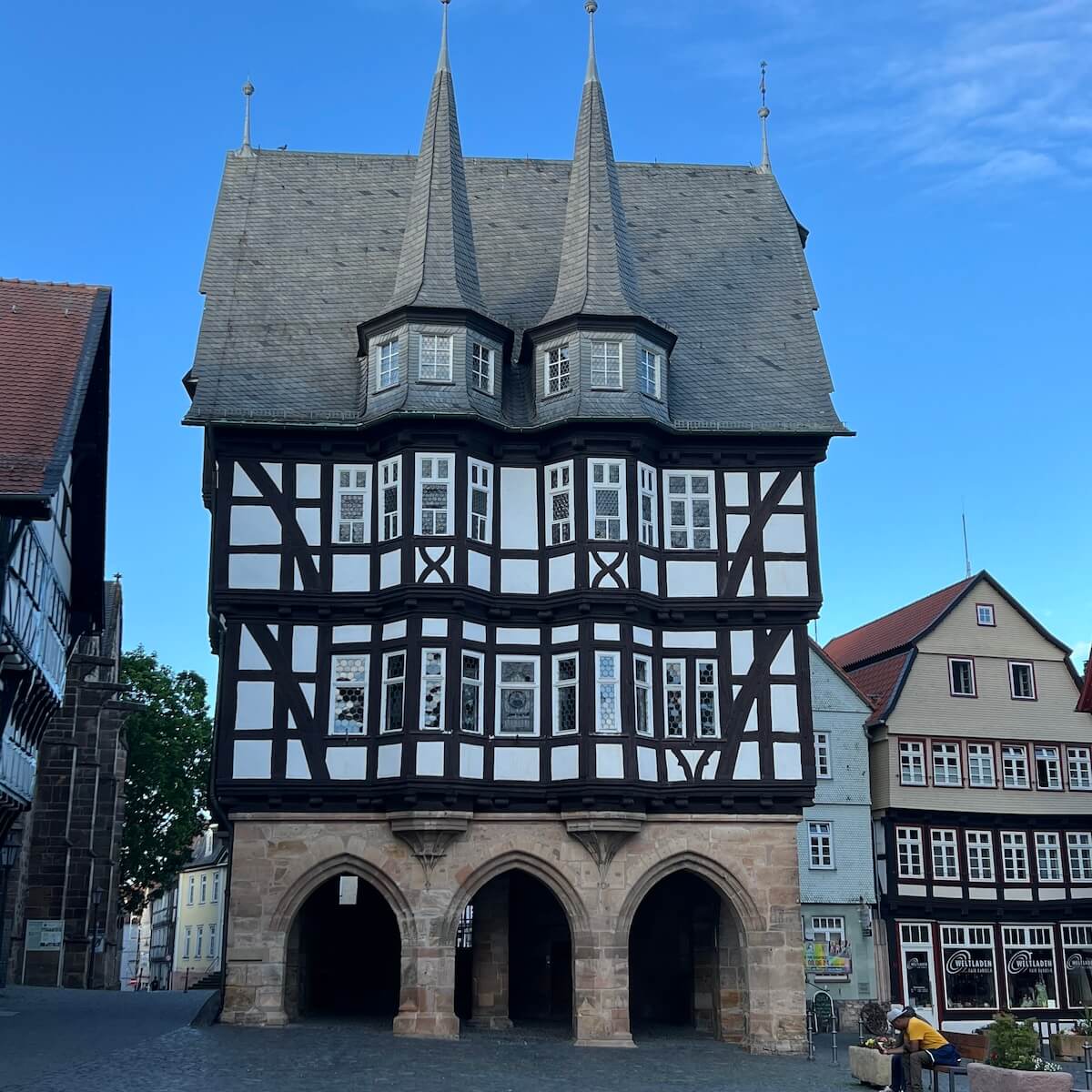 Home 8 Ein großes mittelalterliches Fachwerkgebäude mit zwei hohen, steilen Türmen, einem gewölbten Steinsockel und einer dekorativen Fassade aus weißem und dunklem Holz steht auf dem Alsfelder Marktplatz unter einem strahlend blauen Himmel, umgeben von anderen historischen Gebäuden.