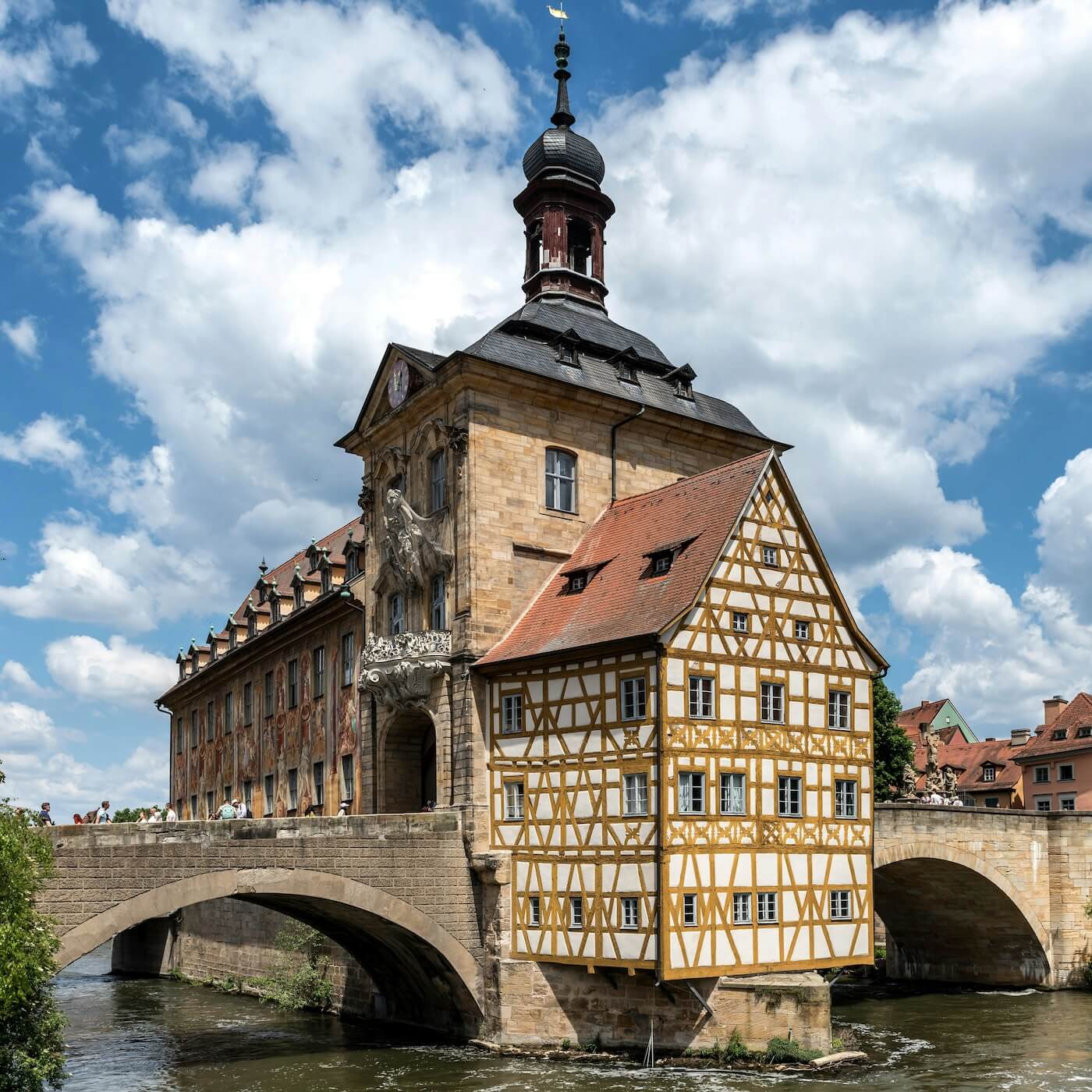 Home 9 Ein historisches Bamberger Gebäude mit Fachwerk und einem Turm steht über einem Fluss, der durch gewölbte Steinbrücken verbunden ist. Das Gebäude weist komplizierte Details und rote Ziegeldächer auf und steht vor einem blauen Himmel mit vereinzelten Wolken. Auf der Brücke sind Menschen zu sehen.