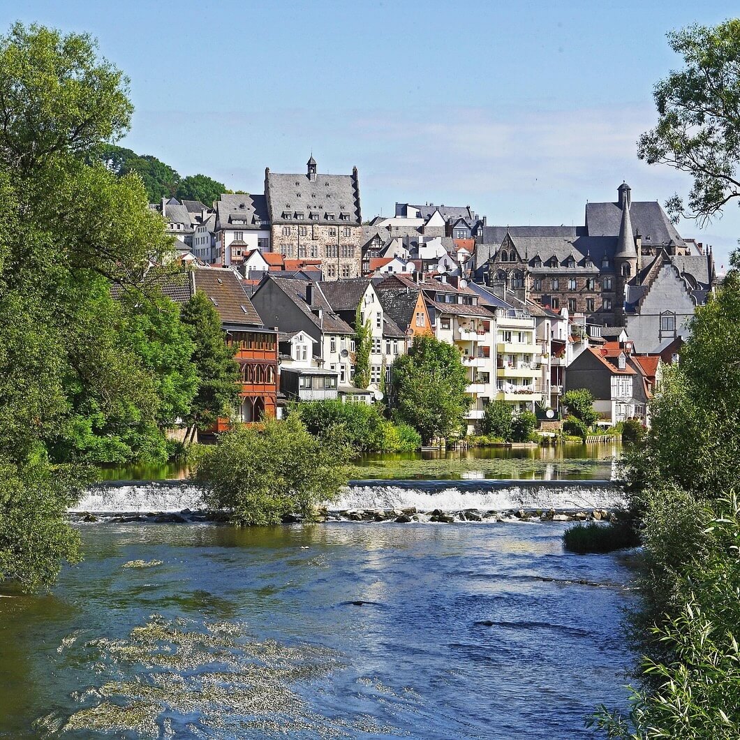 Home 6 Marburg ist eine malerische Stadt am Fluss und bietet historische Gebäude mit spitzen Dächern und eine Mischung aus moderner und alter Architektur. Grüne Bäume rahmen die Szene ein, während im Vordergrund ein sanfter Fluss und ein kleiner Wasserfall unter einem strahlend blauen Himmel fließen.