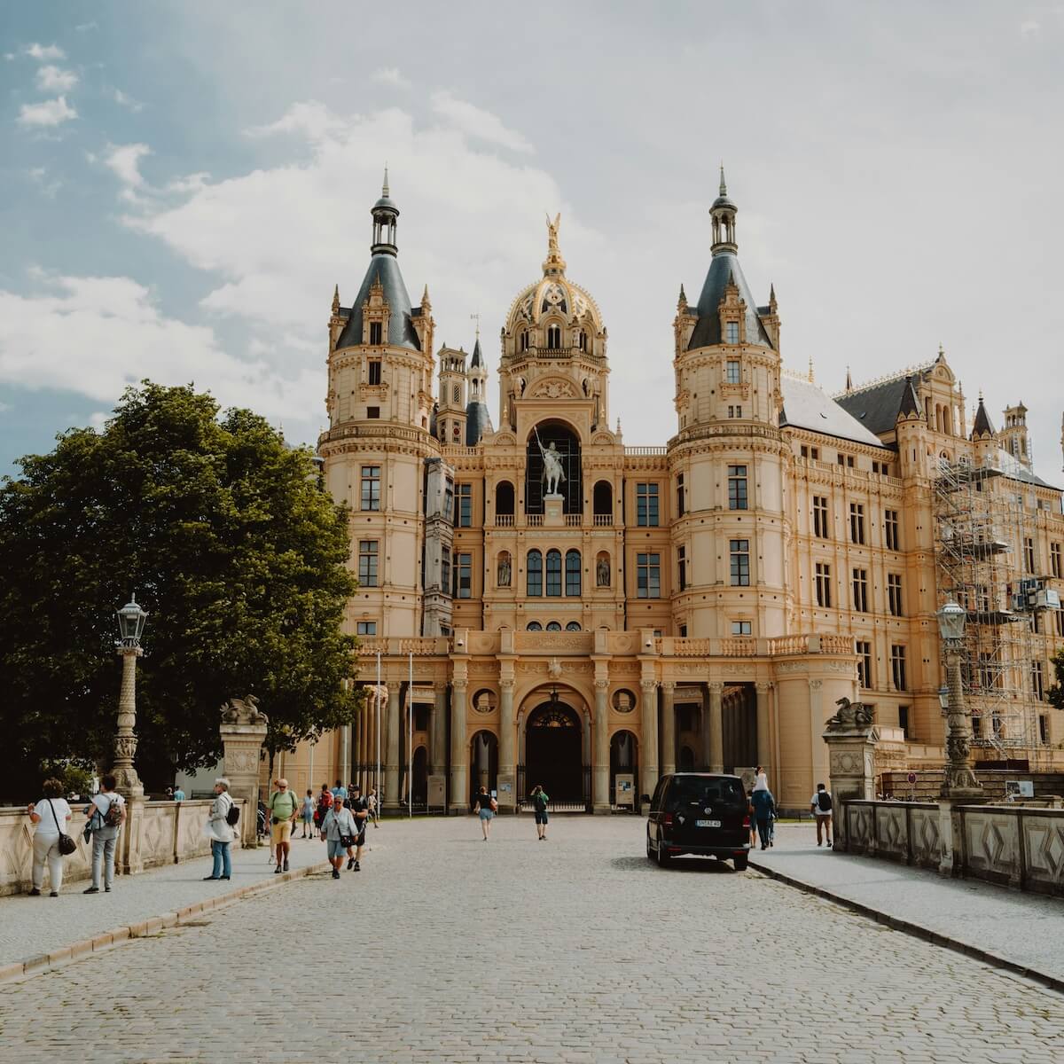 Home 3 Ein prächtiges historisches Schloss mit verschnörkelten Türmen und einer zentralen goldenen Kuppel steht unter einem teilweise bewölkten Himmel in Schwerin. Auf dem kopfsteingepflasterten Weg, der zum Eingang führt, gehen Menschen spazieren und versammeln sich, während Autos und Bäume den Bereich vor dem beeindruckenden Gebäude säumen.